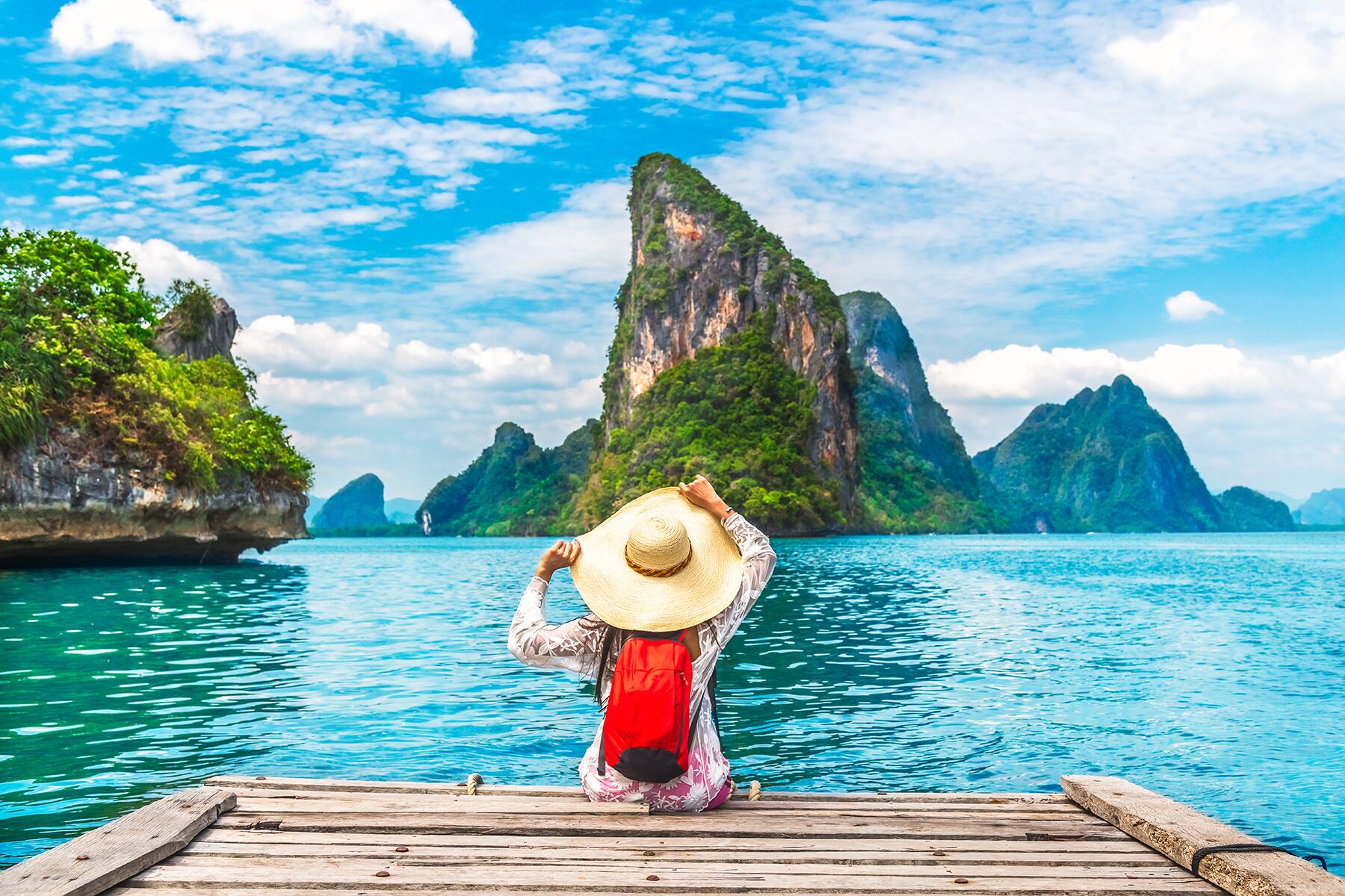 A woman is seated on a dock facing away from the camera, looking out into the ocean at lush rock formations in Phuket, Thailand.