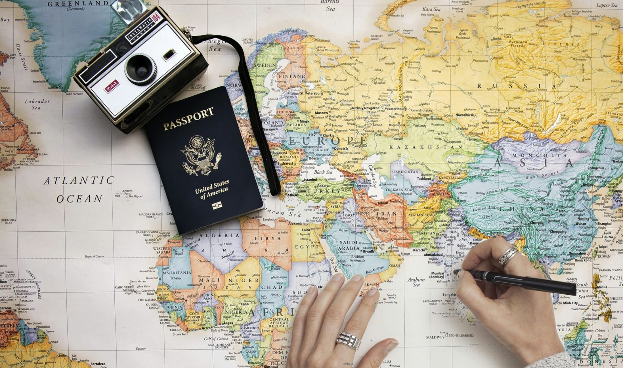 A world map laid out on a table with a USA passport and a film camera on it. A person's hands are visible circling a city and drawing with a marker.